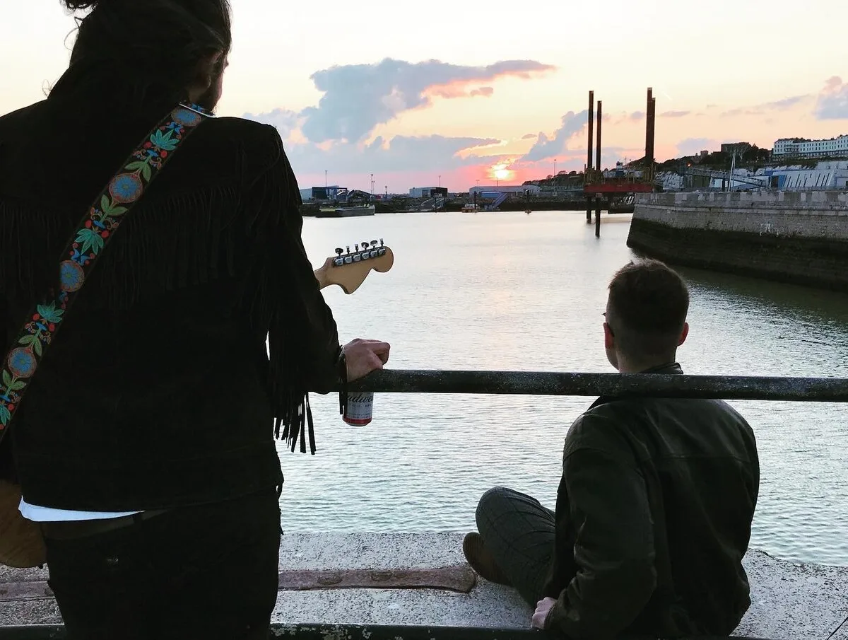 Two musicians at sunset by a harbour, one holding a guitar, looking out over the water