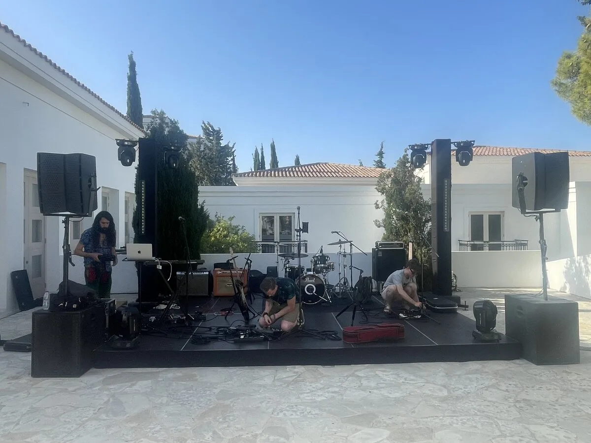 Band setting up on an outdoor stage at a Mediterranean-style venue under blue skies