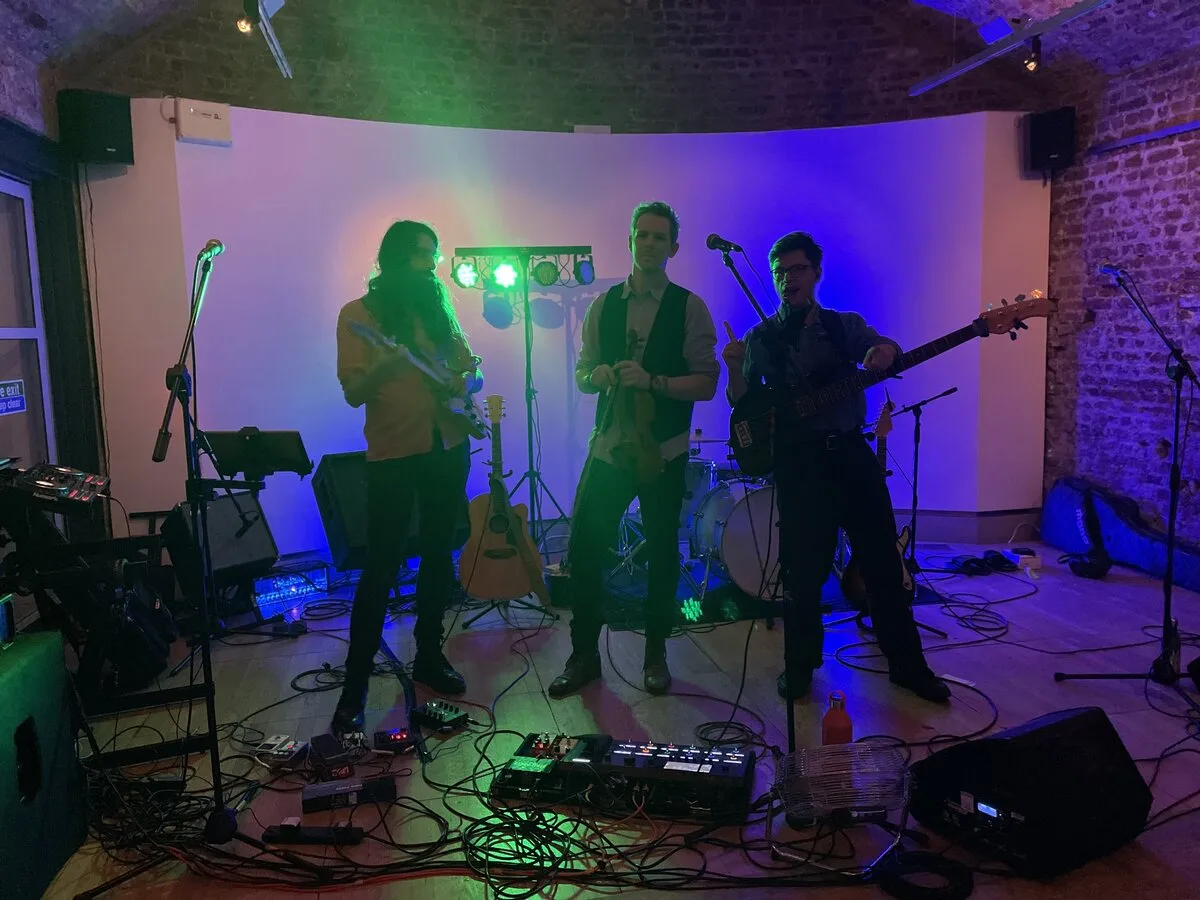 Three band members setting up on stage inside a brick-vaulted venue with colourful stage lighting
