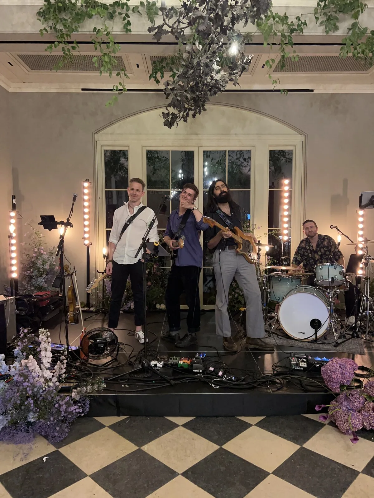 The full band on stage surrounded by floral arrangements, posed before an evening wedding reception